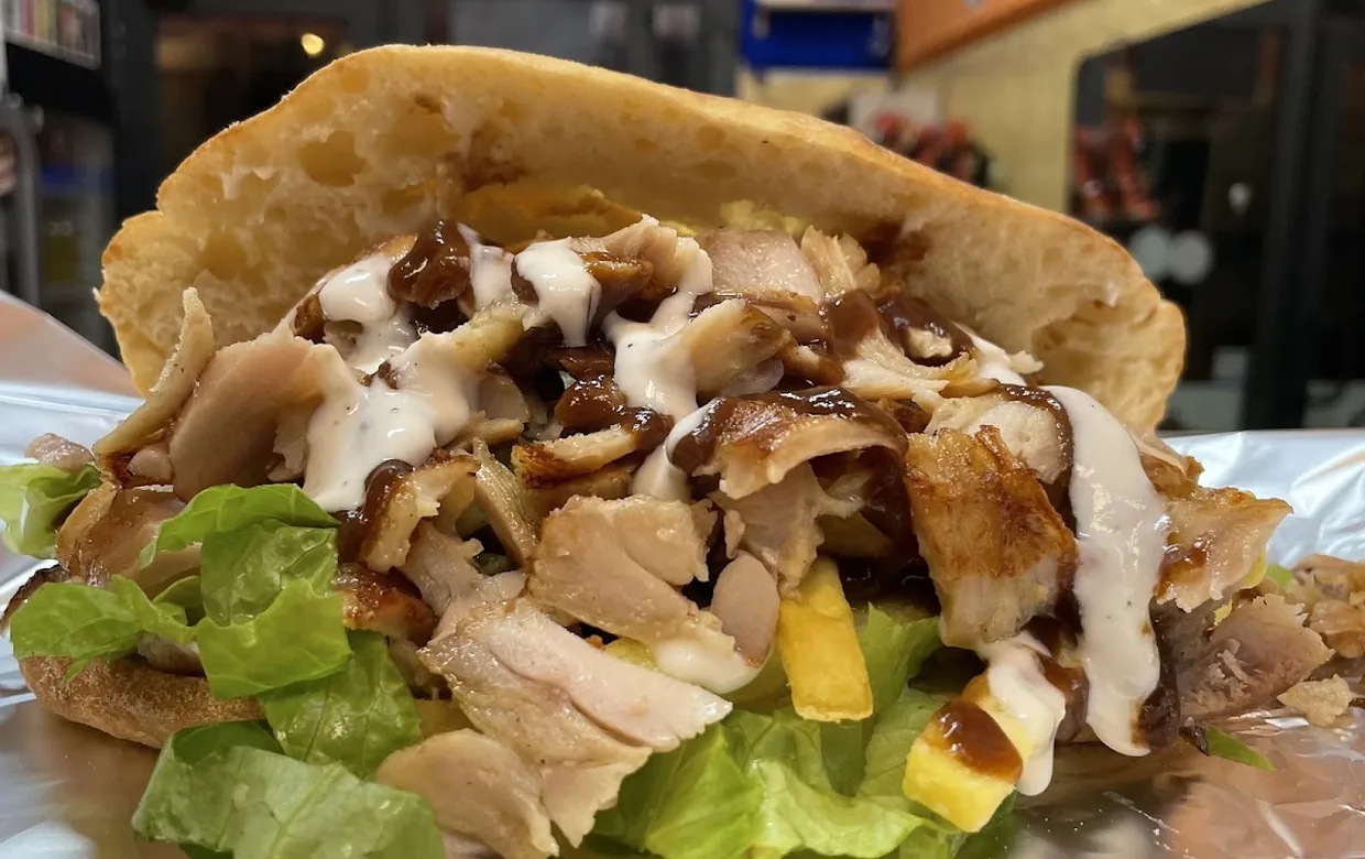 Un panino kebab aperto sul foglio d'alluminio: carne tagliata dallo spiedo, insalata, patatine e salsa bianca, con la piazza fuori dalla porta.